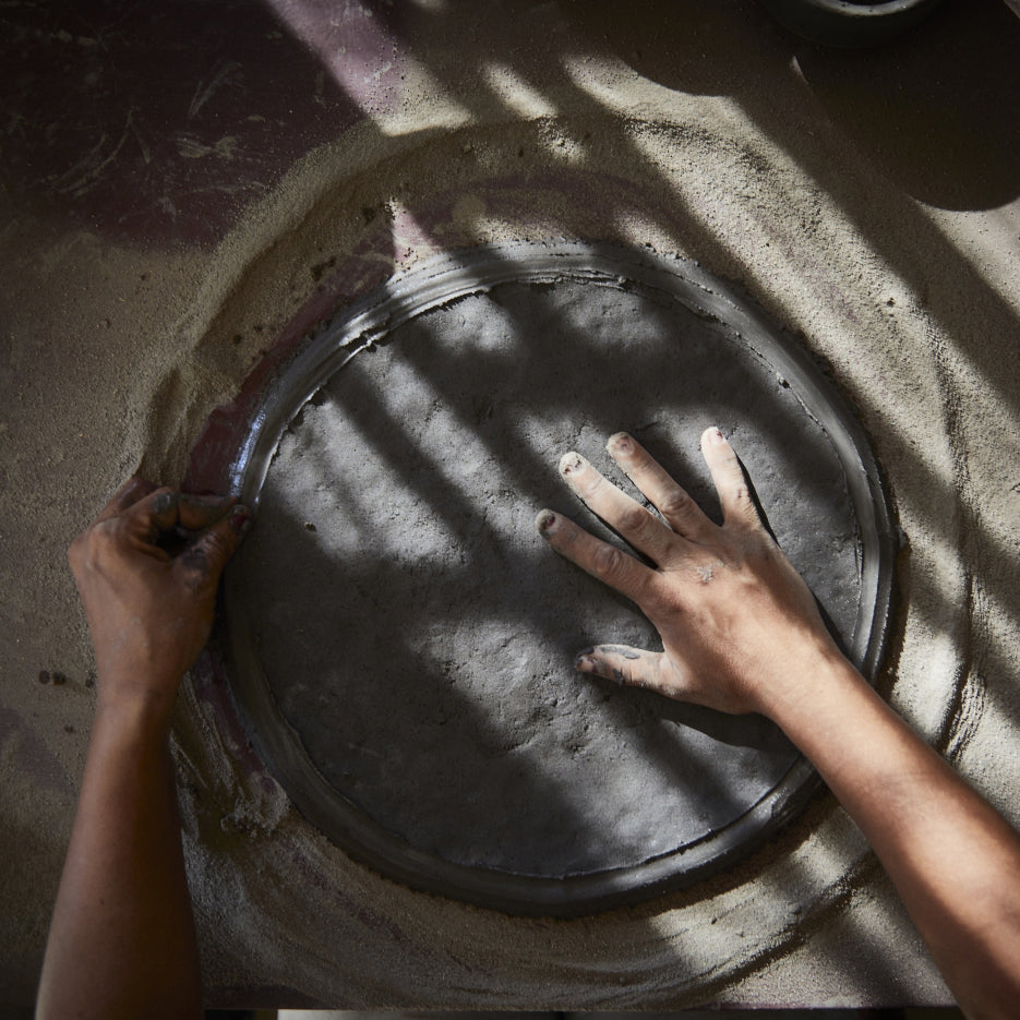 Two hands expertly mold a round clay piece on a sandy surface, echoing traditional Colombian pottery. Sunlight creates striped shadows on the clay and hands, enhancing this artisanal scene as if crafting Ames Loza Ceramics in black.