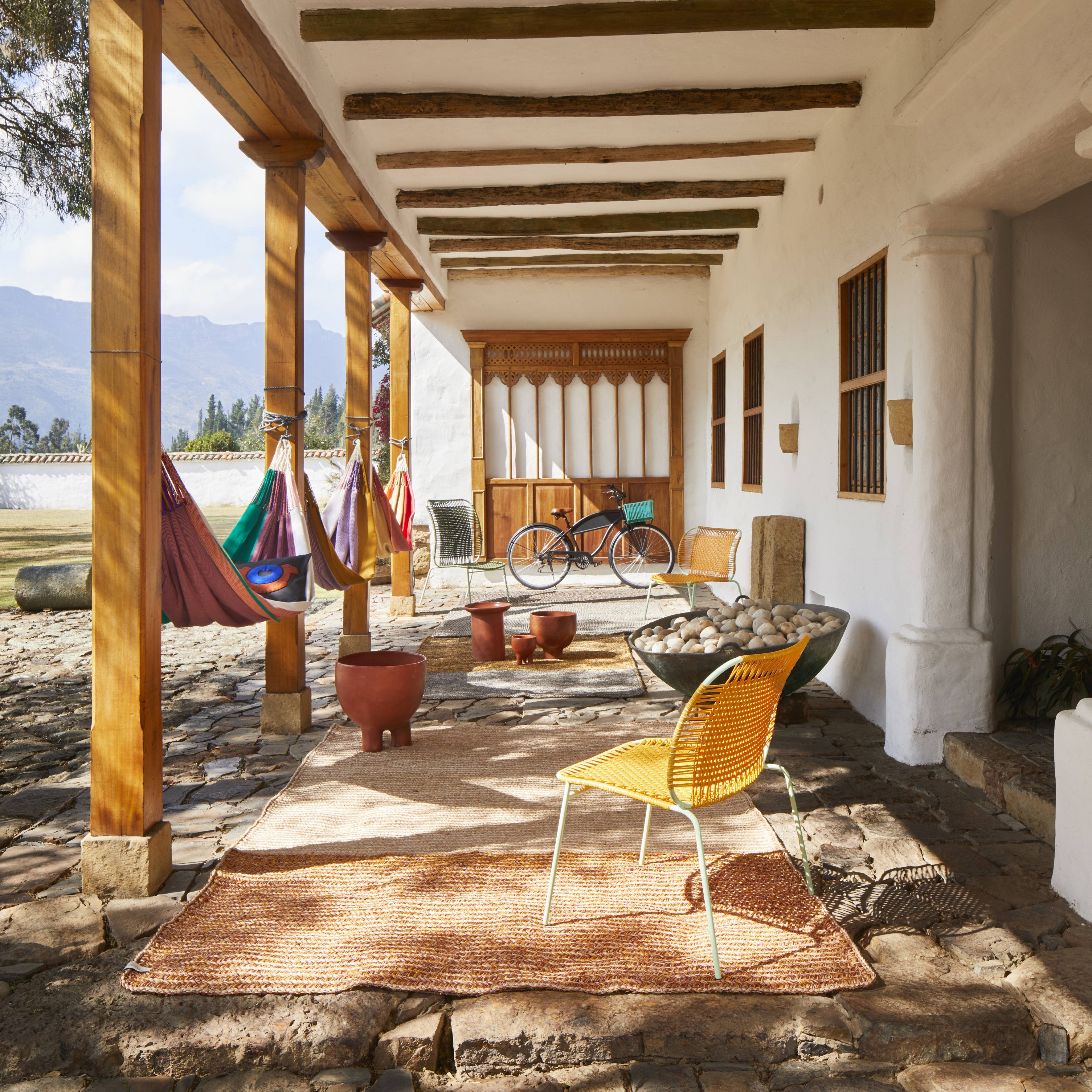 A rustic outdoor veranda features the Ames Cielo Lounge Chair Low by Ames on a woven rug with wooden beams, colorful hammocks, potted plants, and a bicycle by a white stucco wall with mountain views—perfect for indoor or outdoor use.