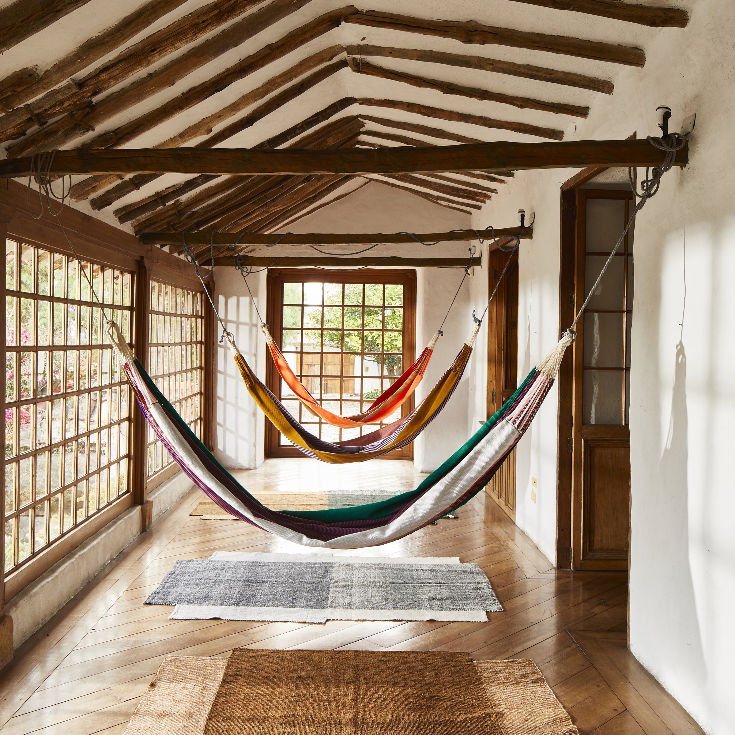 Three Ames Maraca Hammocks in vibrant colors hang in a bright, rustic hallway with wooden beams and large windows. Natural light fills the space, highlighting the wooden floor and creating a cozy, inviting atmosphere with ethnic designs.