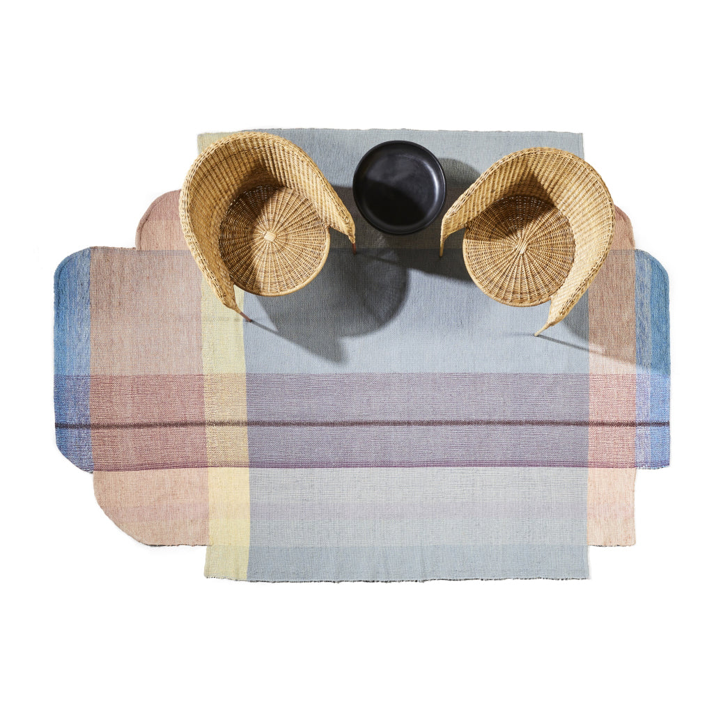 Two wicker chairs and a small black round table are on the Muyska Rug by Ames. The colorful, layered, rectangular hand-woven wool rug features soft pastel shades of blue, pink, and yellow, adding to a cozy contemporary aesthetic. The scene is viewed from above.