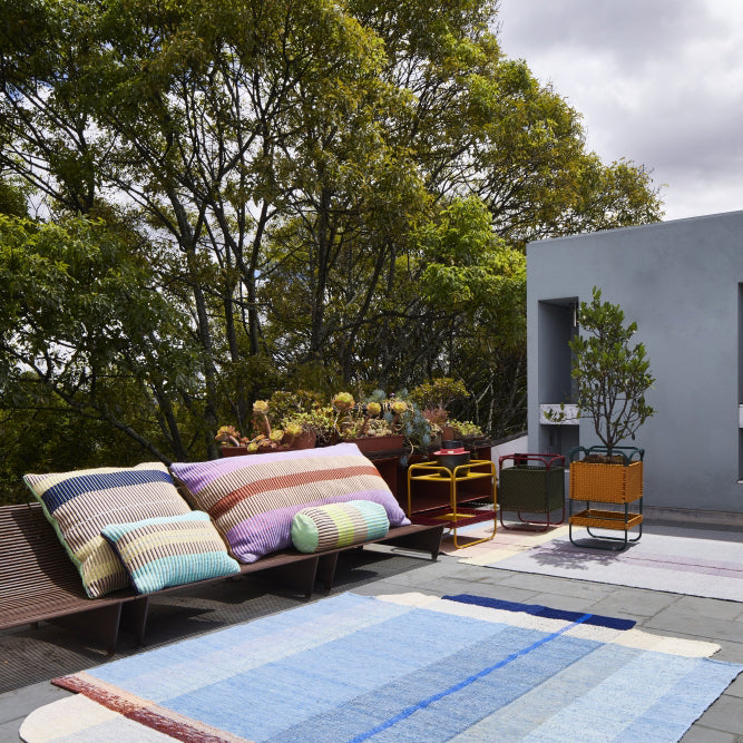 A patio featuring the Ames Muyska Rug by Ames, a modern bench with colorful striped cushions, small tables, and potted plants sits beside a building, surrounded by lush trees under a partly cloudy sky.