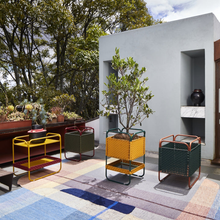 A modern outdoor patio featuring the vibrant geometric Ames Muyska Rug by Ames, square metal planters in yellow, green, and orange, a small tree, potted plants on a table, recessed shelves in a light gray wall, and trees in the background.