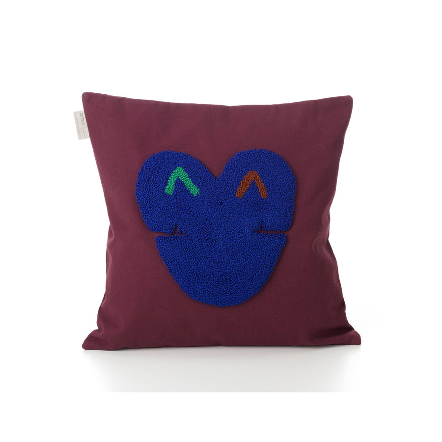 Introducing the Ames Nido Kukuy pillow: a square maroon pillow showcasing a textured blue heart design with abstract green and orange eye-like accents on a plain white 100% cotton background, enhancing its charm.
