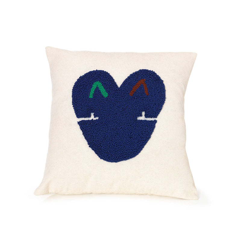 Presenting the Nido Kukuy pillow by Ames: a white square pillow made from 100% cotton, showcasing a textured blue heart. The heart includes two small, colorful arrow-like shapes at the top and minimalist eyes below, infusing charm into any space.