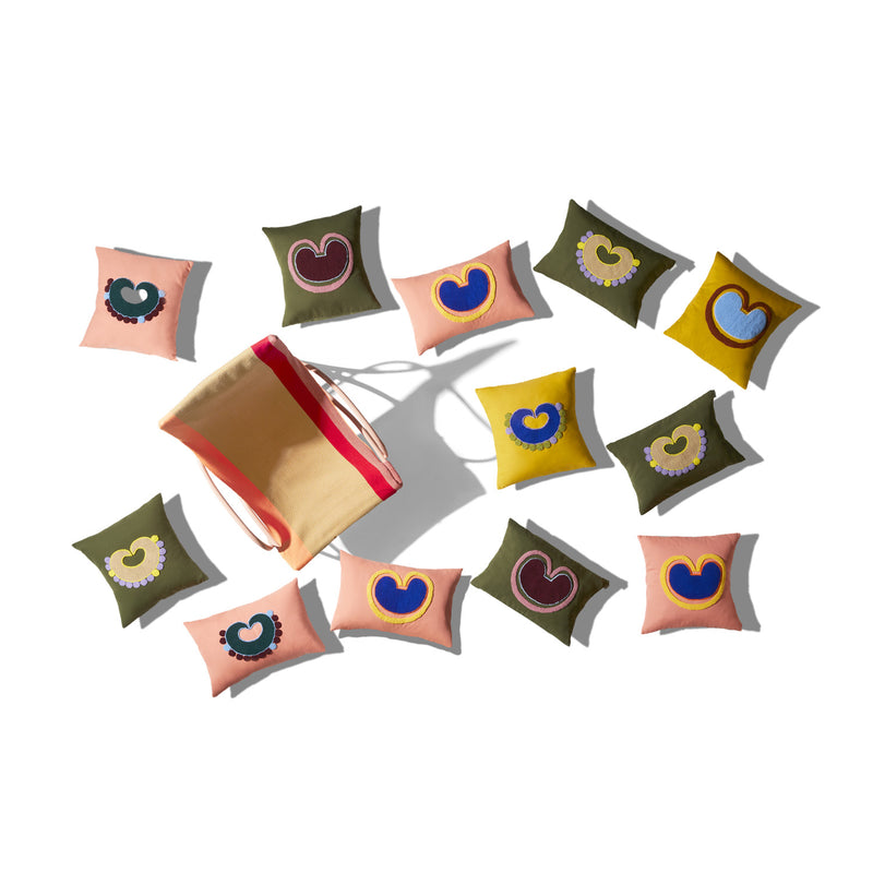 The Ames Nido Coraz collection by Ames features a beige tote bag with red straps, surrounded by green, pink, and yellow handmade cushions decorated with colorful heart appliqués, all set against a white background.