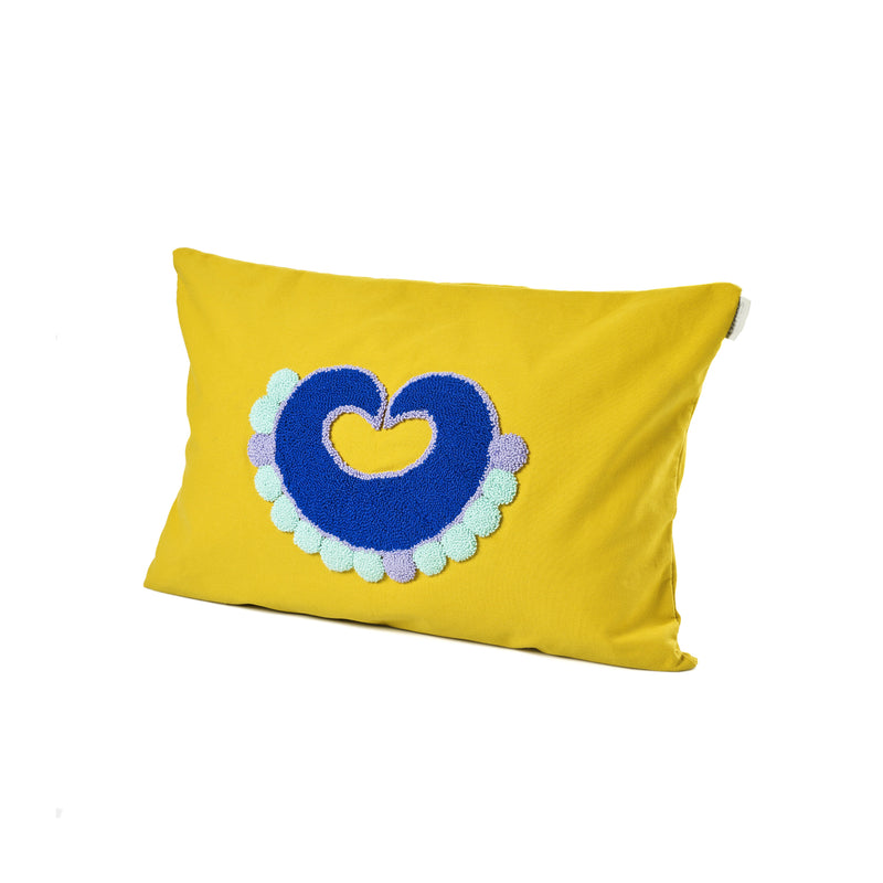 The Nido Piqui cushion by Ames, designed by Sebastian Herkner, features a rectangular yellow pillow with a central blue heart, surrounded by small pastel blue and purple circles. The design draws inspiration from pre-Columbian Tairona motifs.