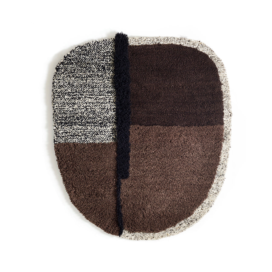 Explore the oval-shaped Nudo Rug by Ames, inspired by Colombian arts and crafts. Its abstract design has four quadrants: speckled black and white in the top left, deep brown on the right, lighter brown at bottom left, and a bold black vertical line anchoring this artistic piece.