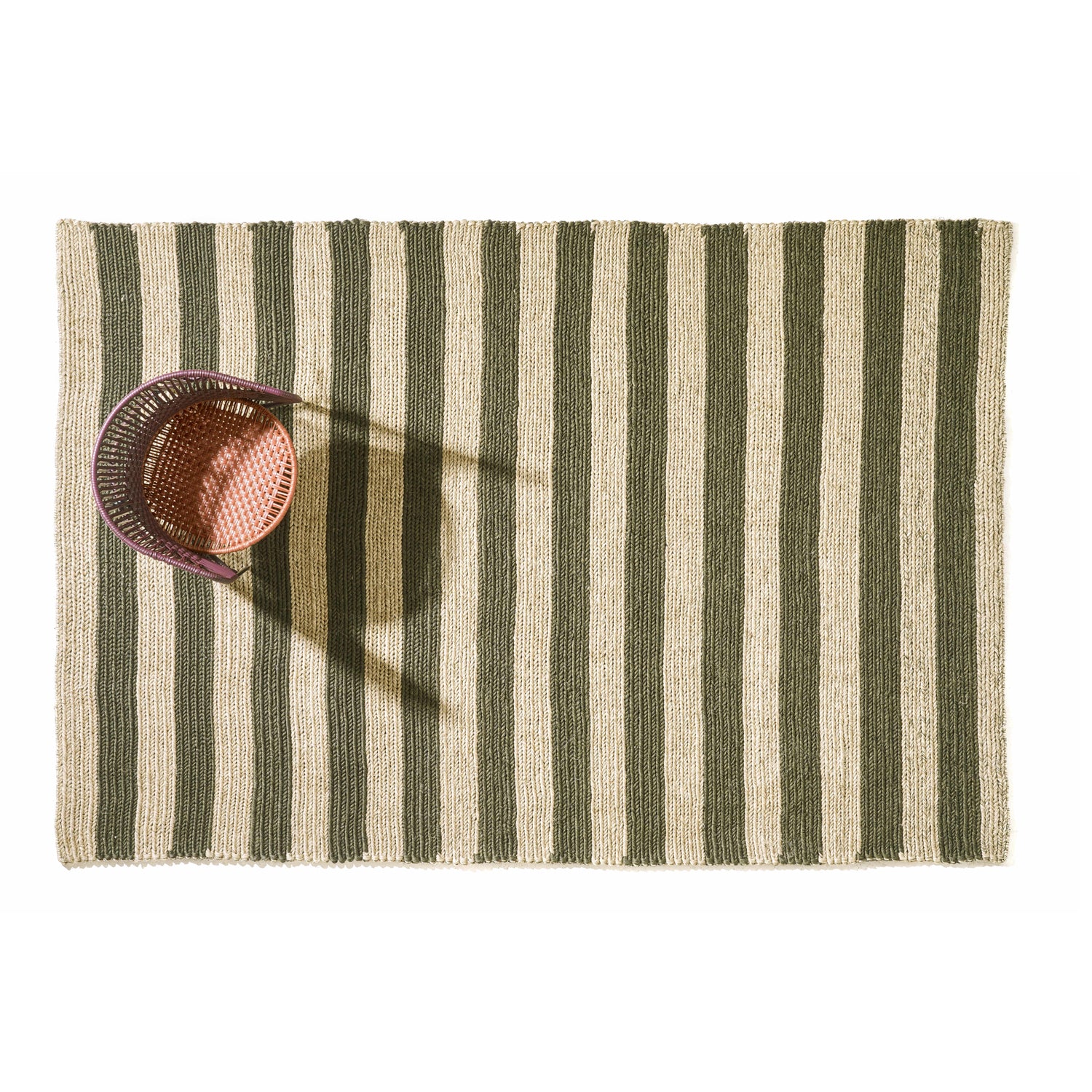 Amess Par Raya Rug features green and beige vertical stripes. From above, a woven basket with a handle casts its shadow onto the rug, located near the top left corner, set against a plain white background that enhances its elegance.