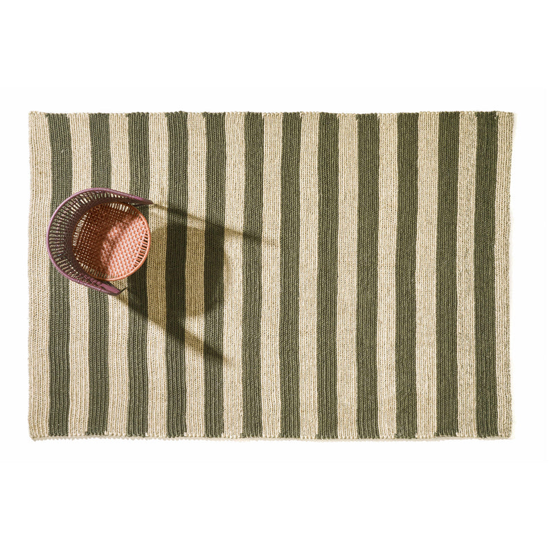 Amess Par Raya Rug features green and beige vertical stripes. From above, a woven basket with a handle casts its shadow onto the rug, located near the top left corner, set against a plain white background that enhances its elegance.