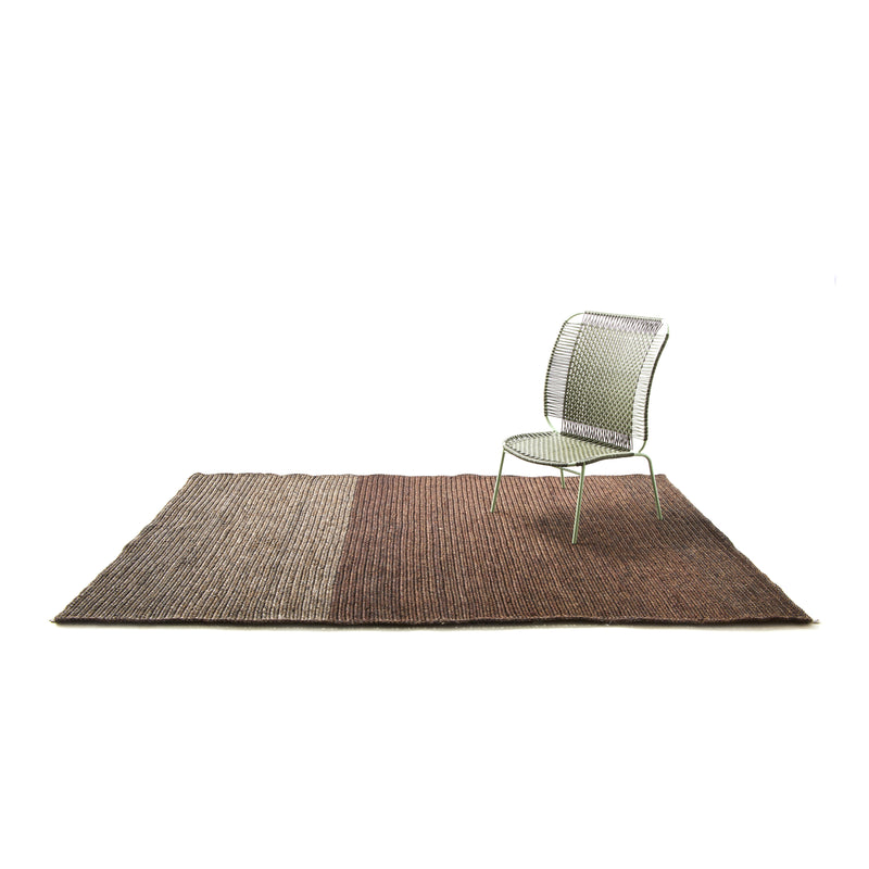 A metal mesh chair sits on the Ames Par Rug by Ames, a rectangular, two-tone brown handmade rug crafted from fique fibers, set against a plain white background.