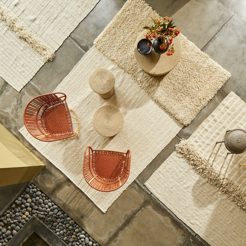 Overhead view of a modern living space featuring the beige textured Ames Subas Rug by Ames, two round woven tables, orange wicker chairs, a vase with flowers, and smooth stone accents along a concrete floor.
