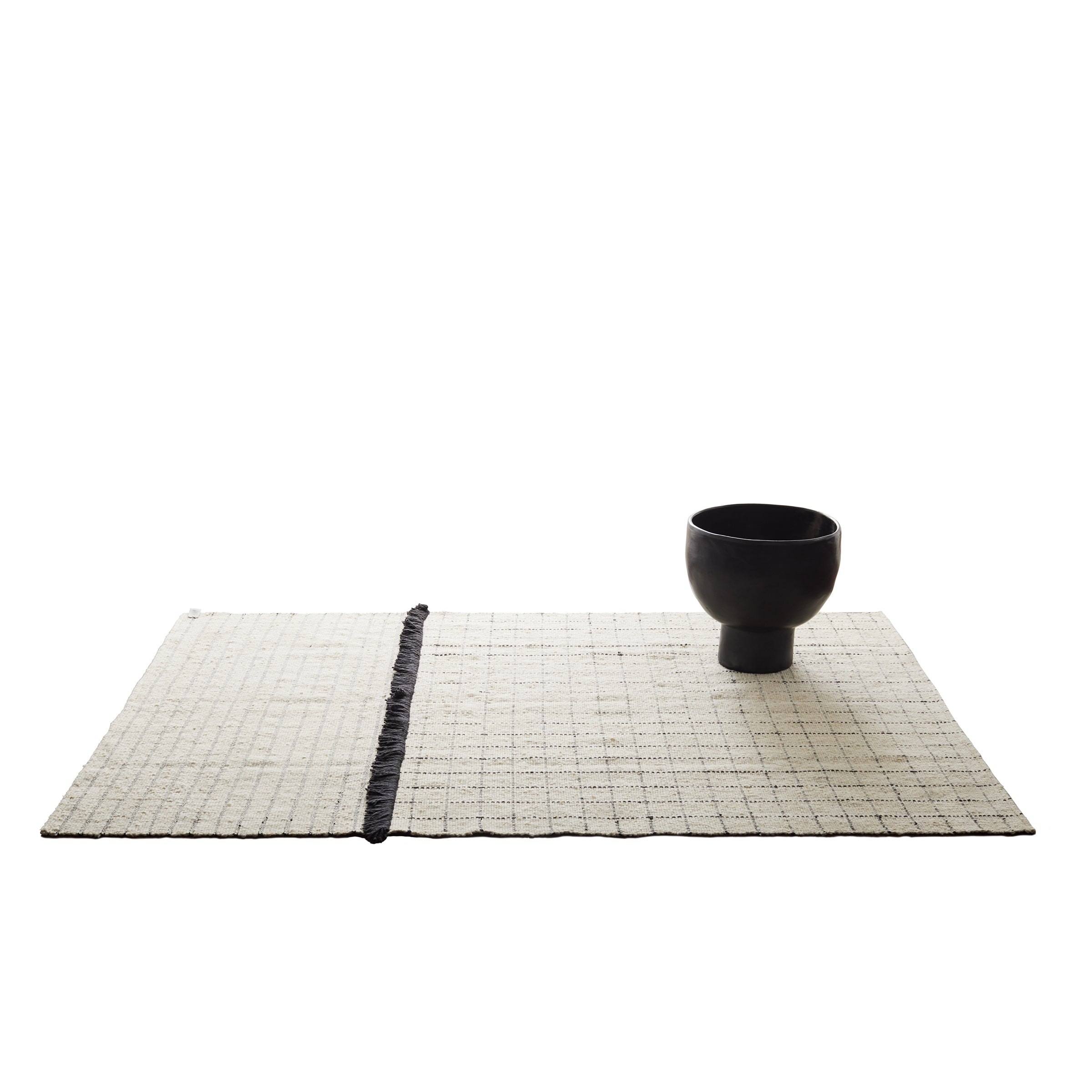 A black ceramic bowl sits on the right side of a rectangular, light-colored woven mat with a dark border stripe, resembling the Ames Subas Rug by Ames, both arranged on a white background.