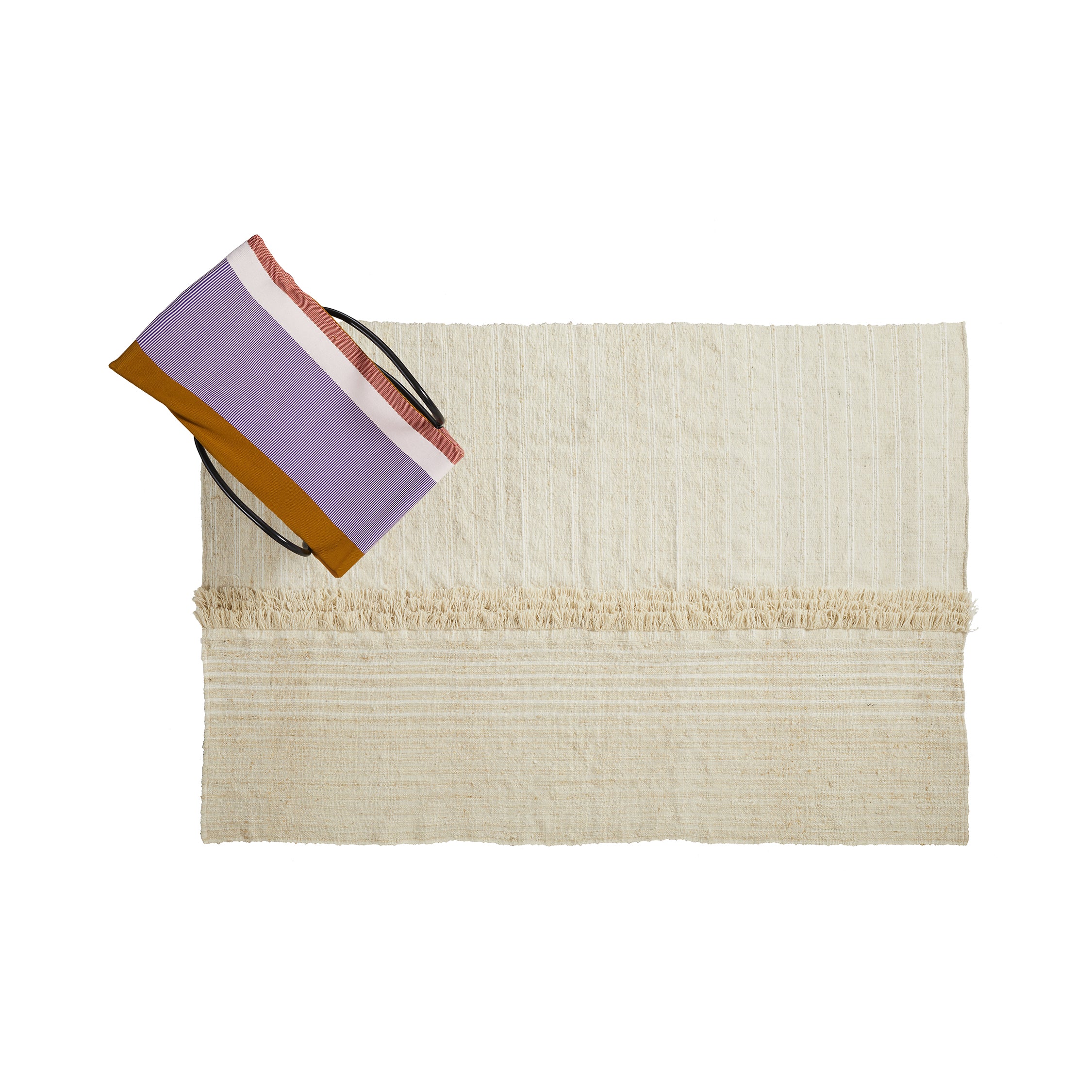A folded cream-colored Ames Subas Rug by Ames is placed next to a striped tote bag in purple, pink, mustard yellow, and white against a white background.