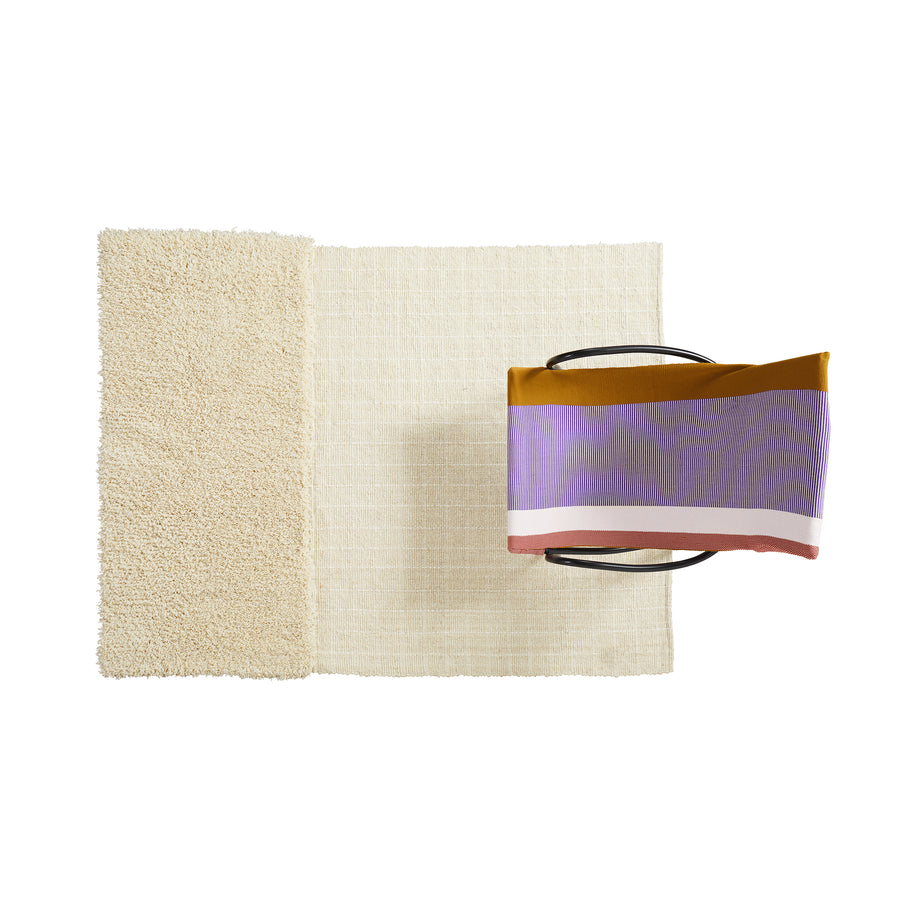 Overhead view of a modern chair with a purple, mustard yellow, and pink striped cushion, placed on two overlapping Ames Subas Rugs by Ames. The rugs are cream-colored wool on a white background. Design inspired by Sebastian Herkner.