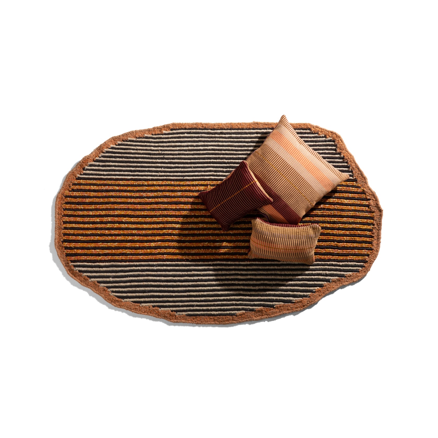The Ames Uilas Noche Rug, featuring orange and black stripes made from dyed black fique fiber, lies on a white background. Three textured pillows in brown and tan shades are placed on the rug, creating a cozy luxurious arrangement.