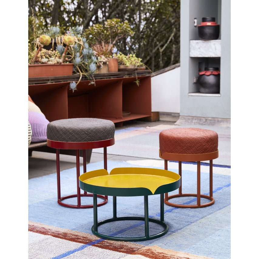 Three modern, round Ames Victorias Poufs with textured upholstery surround a small yellow and green Amazonia coffee table on a patio, accented by plants and shelves in the background.