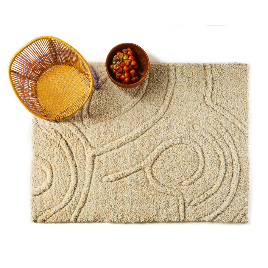 A beige Zenues Rug by Ames, featuring Zenu-inspired circular patterns, lies beside a wicker basket. Close by, a brown bowl filled with small tomatoes enhances the serene display on a white backdrop.