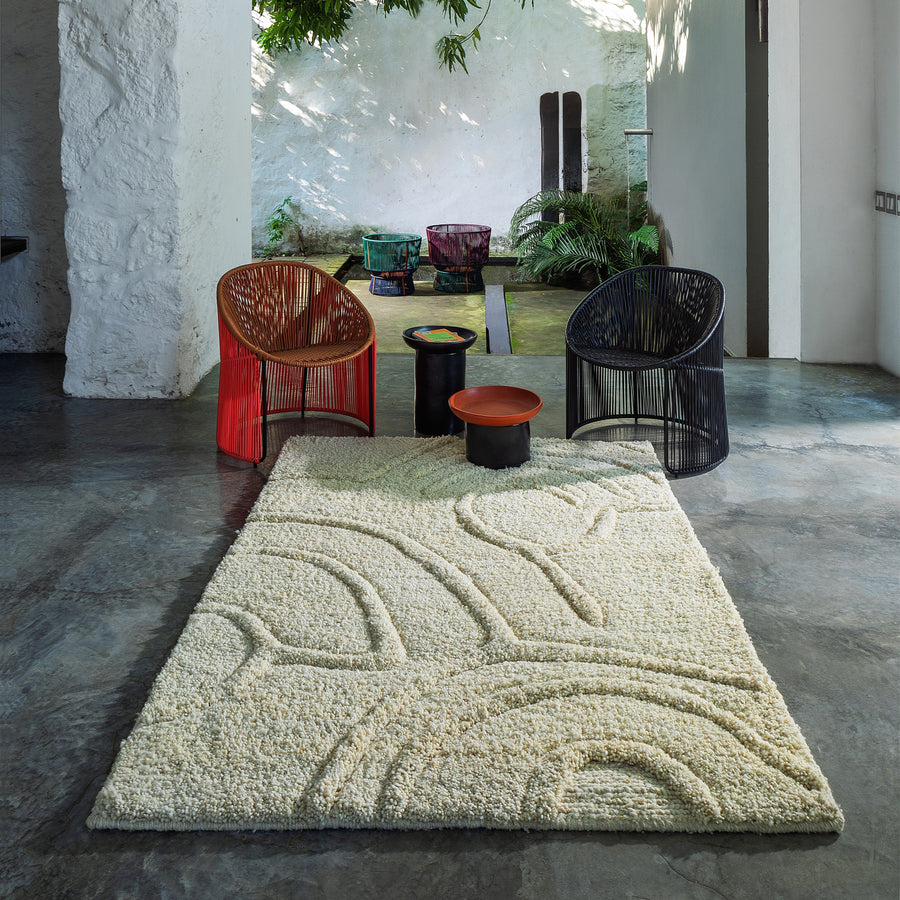A modern living space showcases a cream rug with abstract designs similar to the Zenues Rug by Ames. Two woven chairs, one red and one black, hint at Sebastian Herkners style, arranged around two small side tables. An open doorway reveals a lush outdoor garden in the background.