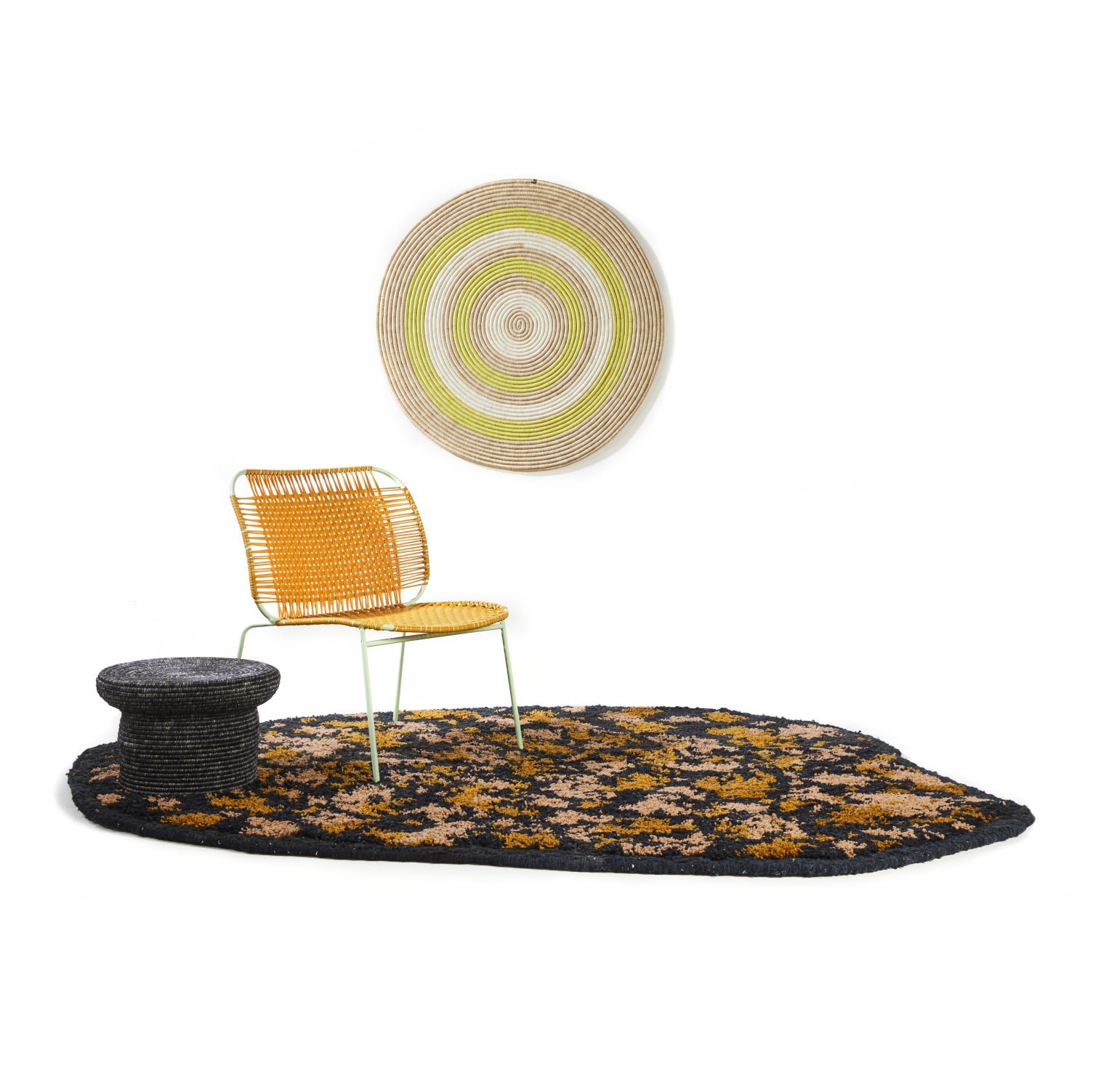 A modern seating area featuring a yellow woven chair, a round black side table, the Ames Zipa Rug by Ames in black, gold, and brown, and a round wall hanging with beige and yellow concentric circles.