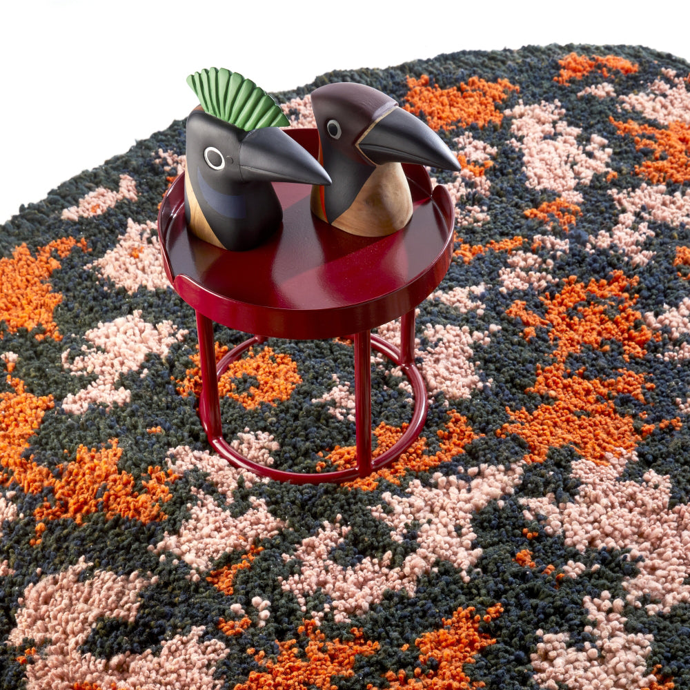 A small round red table displays two decorative wooden bird figurines on the Ames Zipa Rug by Ames, a vibrant piece with abstract black, orange, and pink patterns.