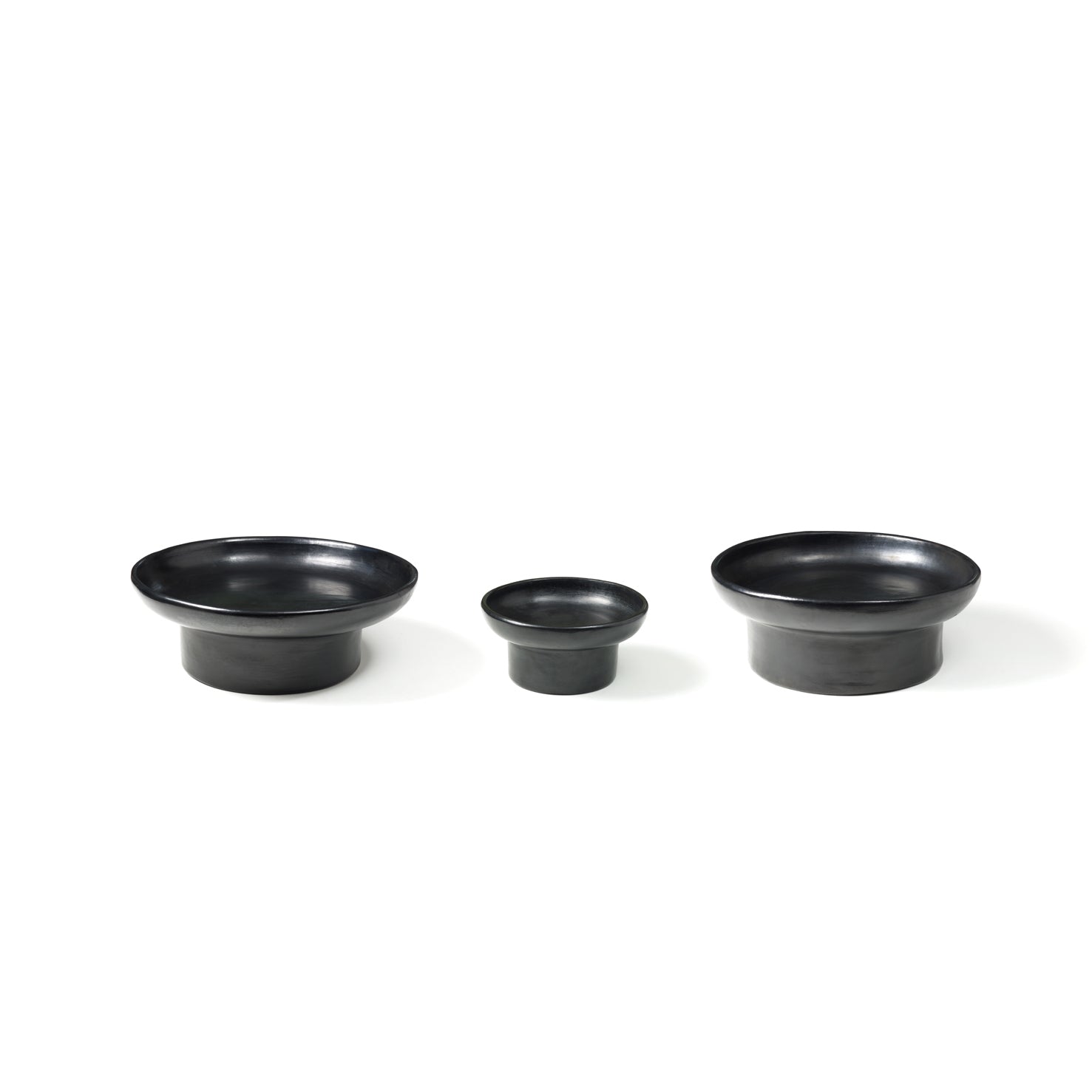 Arranged in a row against a white background, the Barro Tray 1 by Ames features three black ceramic bowls of different sizes. Showcasing traditional pottery techniques, these bowls have a smooth matte finish and slightly flared rims, with the smallest bowl in the center.