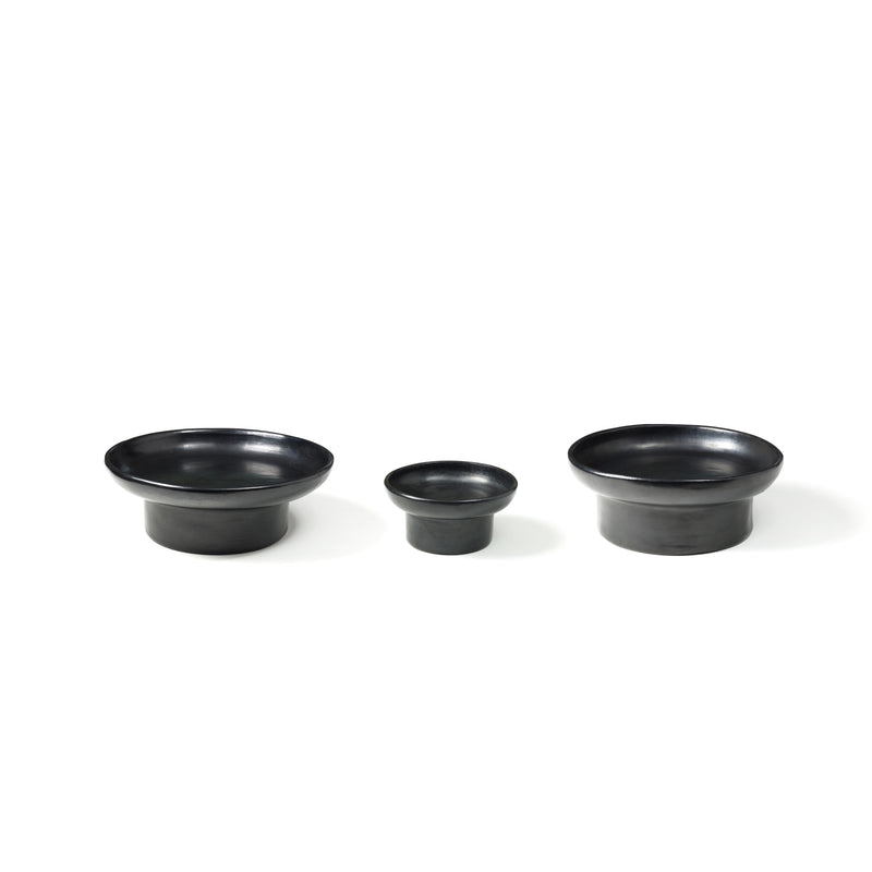 Arranged in a row against a white background, the Barro Tray 1 by Ames features three black ceramic bowls of different sizes. Showcasing traditional pottery techniques, these bowls have a smooth matte finish and slightly flared rims, with the smallest bowl in the center.