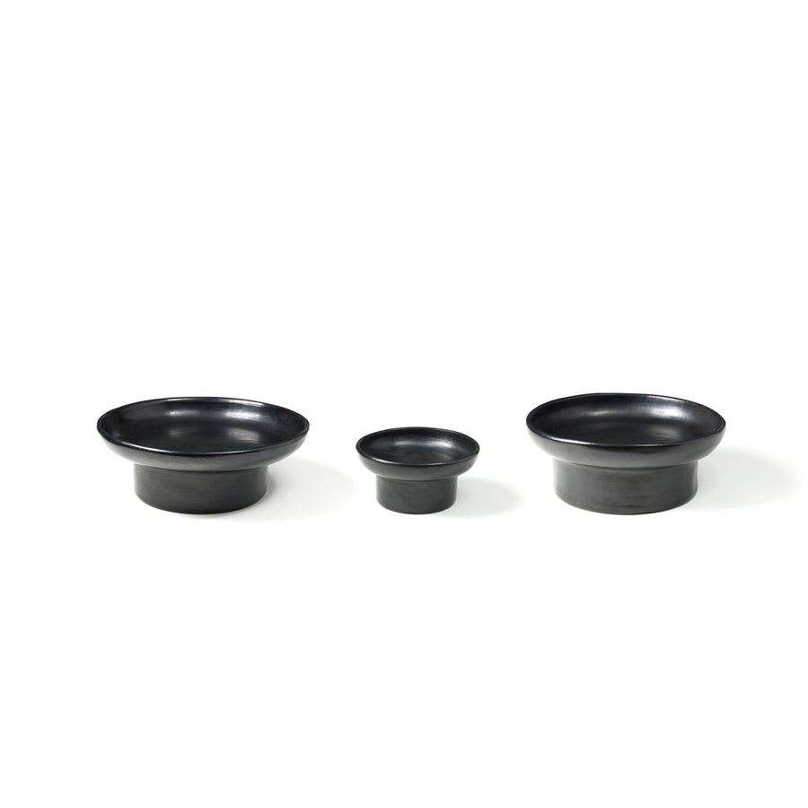 Arranged in a row against a white background, the Barro Tray 1 by Ames features three black ceramic bowls of different sizes. Showcasing traditional pottery techniques, these bowls have a smooth matte finish and slightly flared rims, with the smallest bowl in the center.