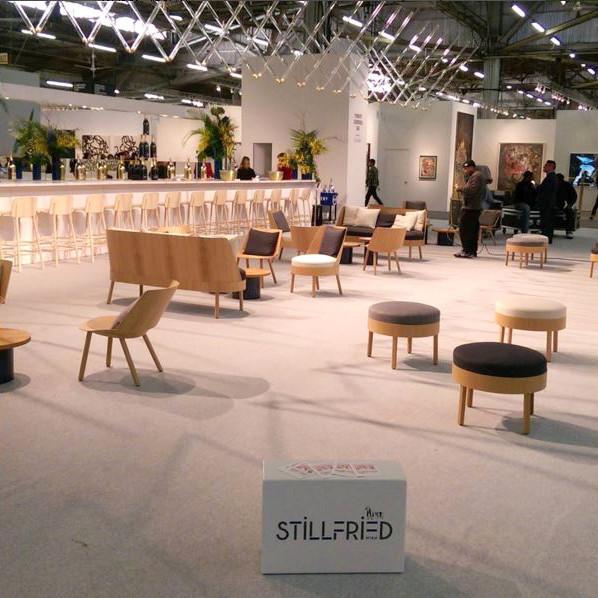 The indoor lounge features modern wooden EC02 BESSY Lounge Chairs by E15. A long bar, oak-veneered plywood tables, and plants enhance the well-lit space. A prominent white STILLFRIED box adds to the decor.