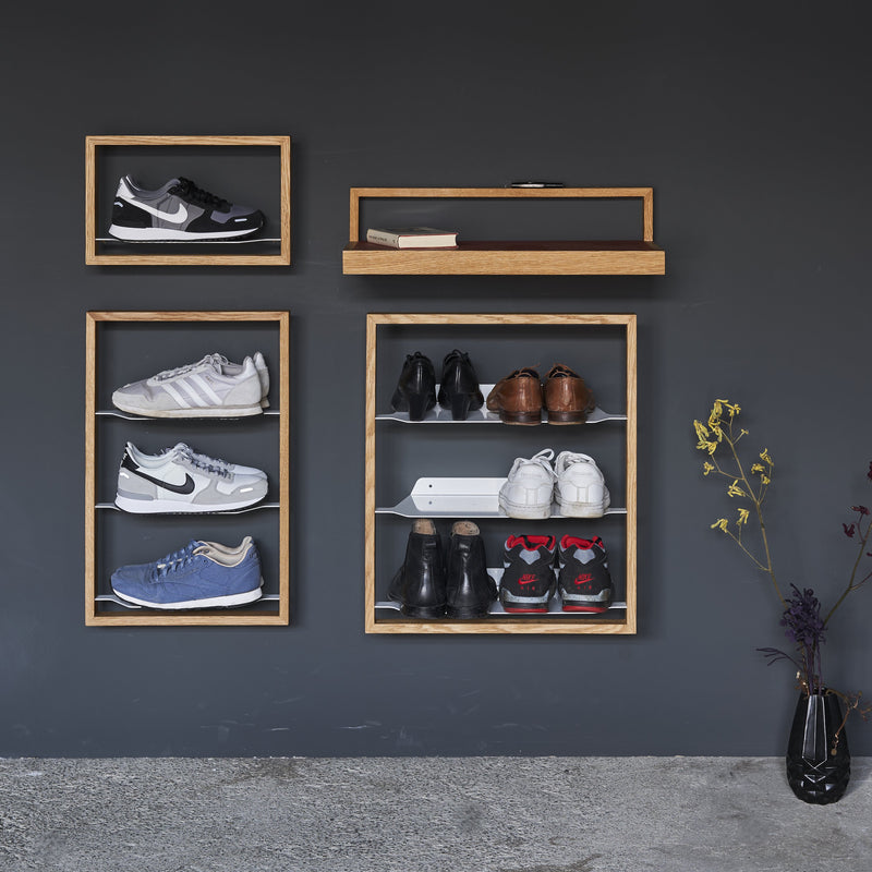 The Basti - Presentation by Das kleine b is a wall-mounted wooden shoe rack displaying sneakers and shoes against a gray backdrop. It offers framed sections for display, includes a small shelf, and is complemented by an artful vase with dried flowers on the ground.