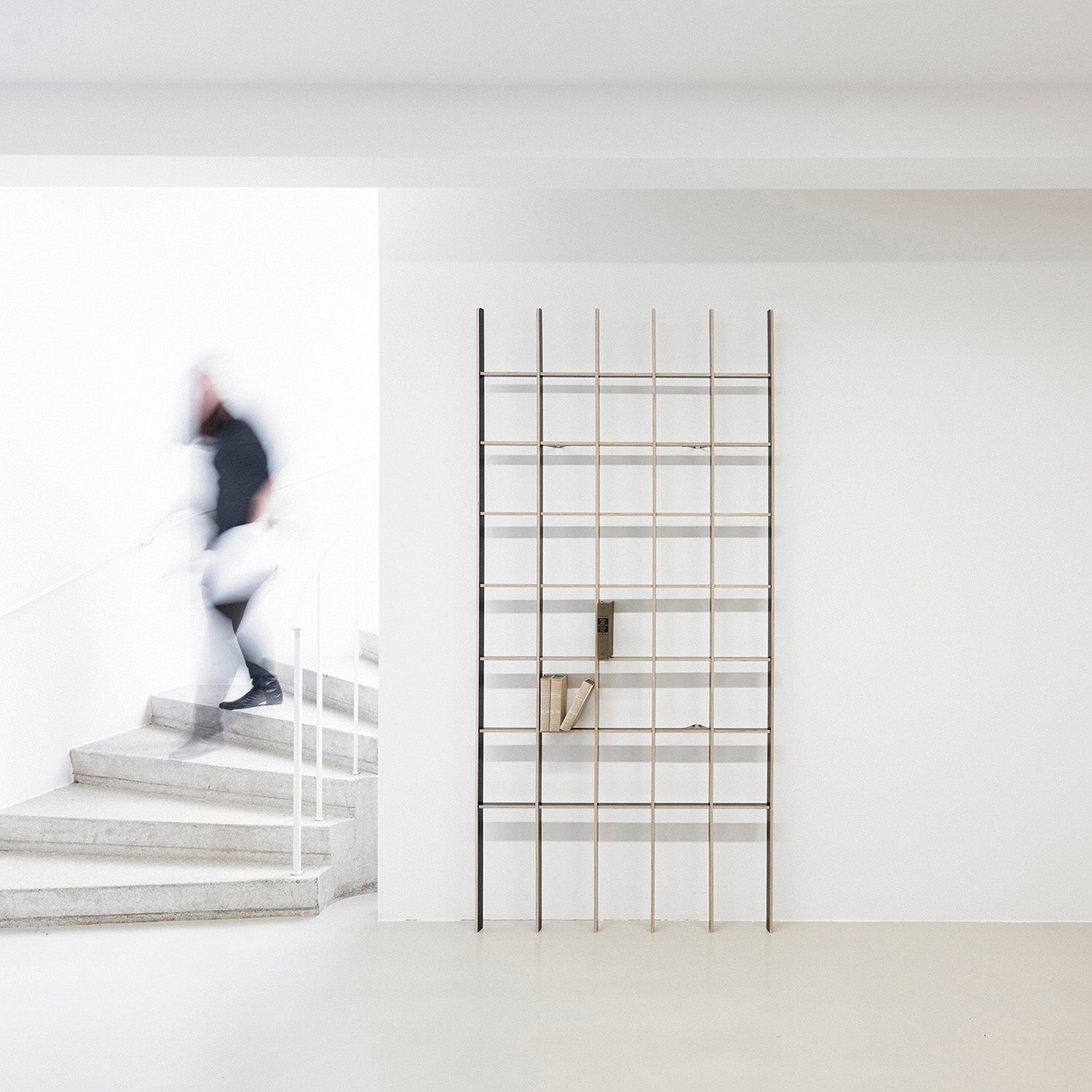 A person in motion descends a minimalist grey staircase beside the Open metal grid bookcase by Martin Breuer Bono, which complements the design with a small sculpture and book on display against a white wall, fostering a modern and artistic atmosphere.