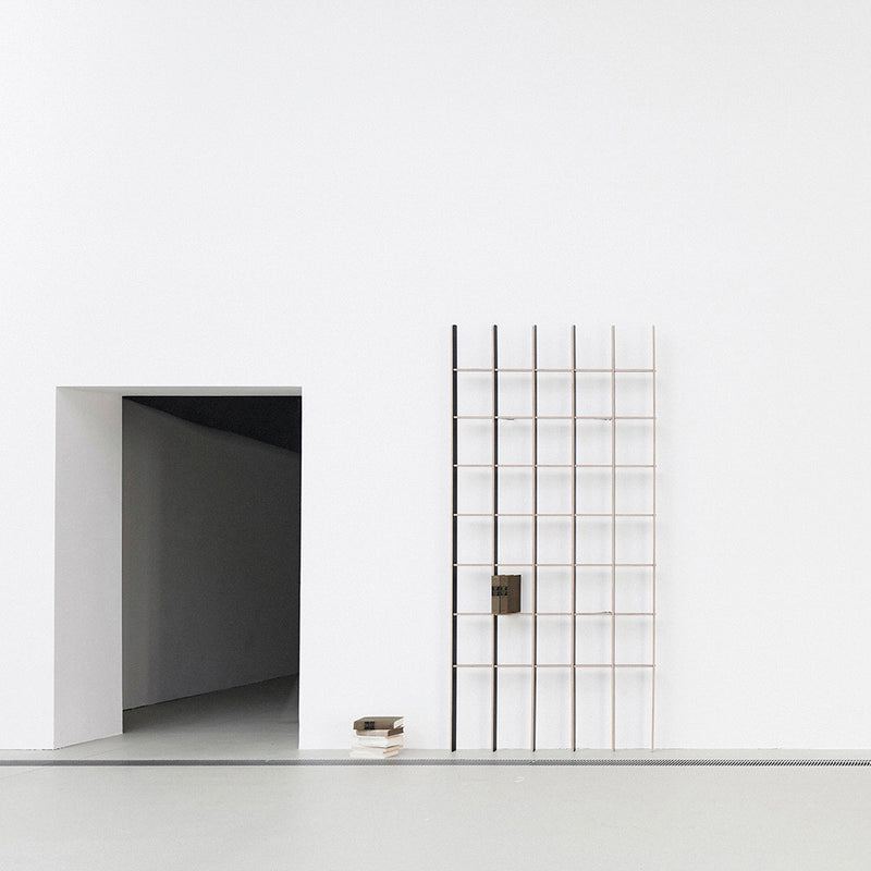 A minimalist room features a large white wall. To the left, a rectangular opening leads to a hallway. On the right, the Martin Breuer Bono Open, a modern grid-style shelving unit, is mounted on the wall, embodying sleek design with a small box and stacked books neatly on the floor.
