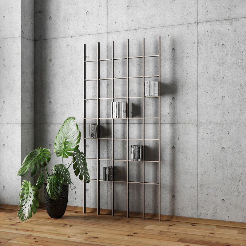 The Open bookshelf by Martin Breuer Bono, with its minimalist grid design, stands against a concrete wall. Made from plywood, it neatly holds several books. A large potted monstera on the left adds greenery, enhancing the sleek aesthetic against the wooden floor.