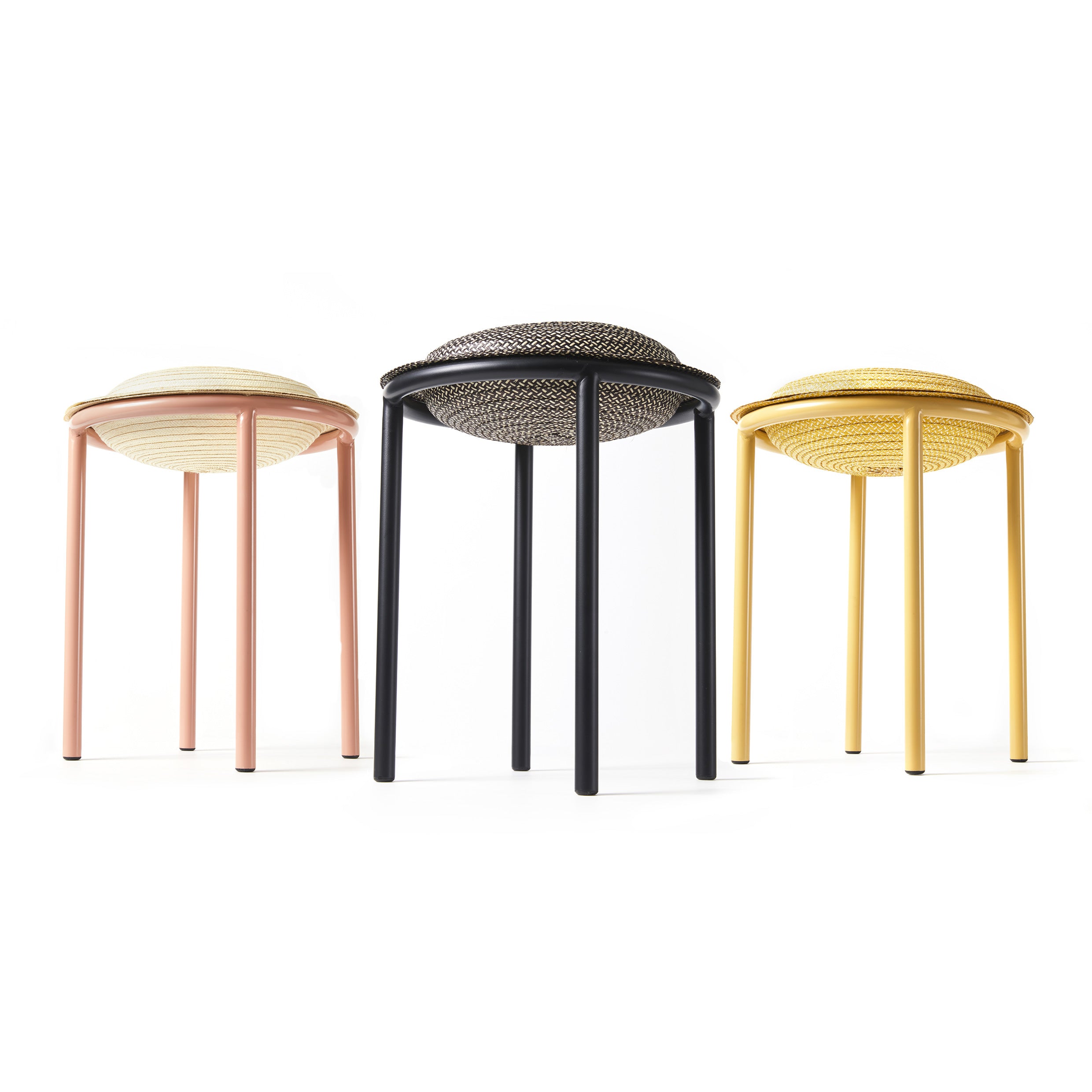 The Ames Cana Stools feature three vibrant designs in pastel pink, black, and yellow. With circular seats and four slender legs each, these stools have woven patterns that stand out against a plain white background, highlighting their unconventional style.