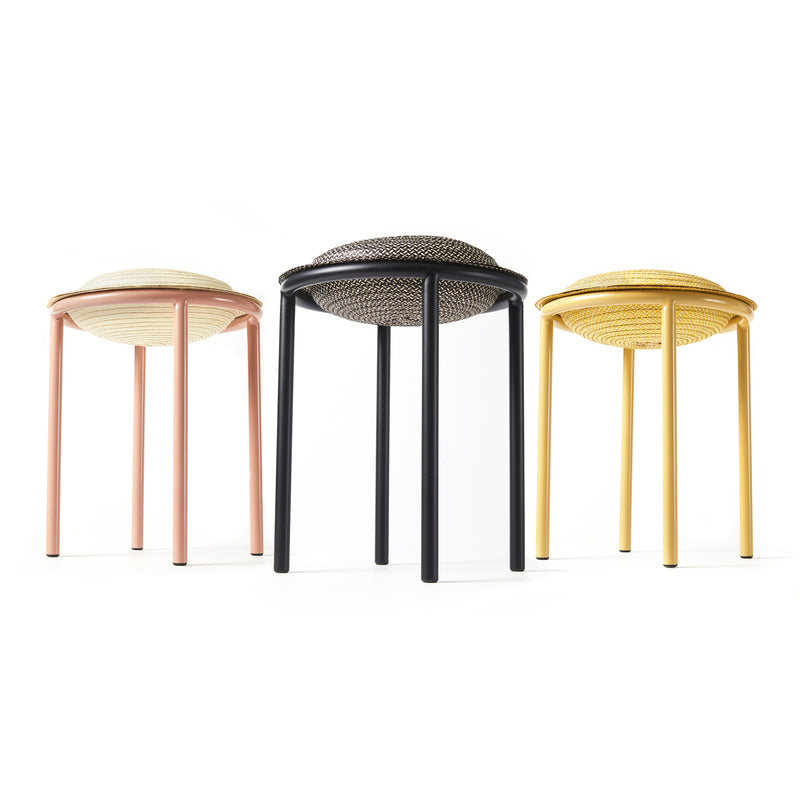 The Ames Cana Stools feature three vibrant designs in pastel pink, black, and yellow. With circular seats and four slender legs each, these stools have woven patterns that stand out against a plain white background, highlighting their unconventional style.