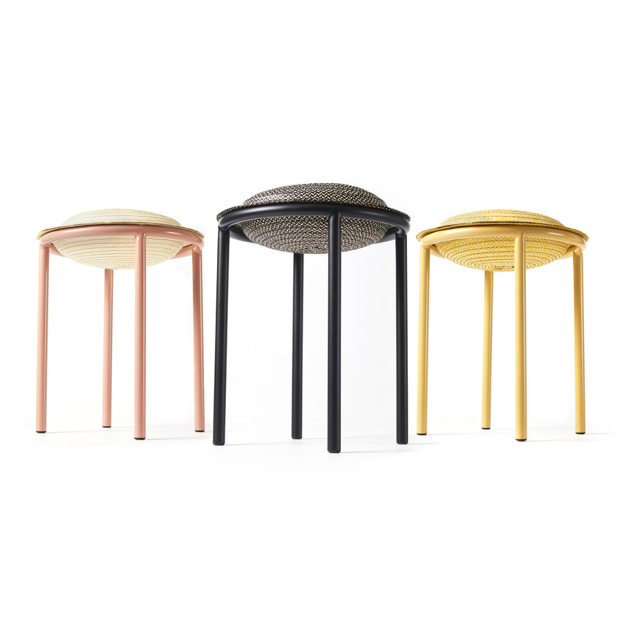 The Ames Cana Stools feature three vibrant designs in pastel pink, black, and yellow. With circular seats and four slender legs each, these stools have woven patterns that stand out against a plain white background, highlighting their unconventional style.