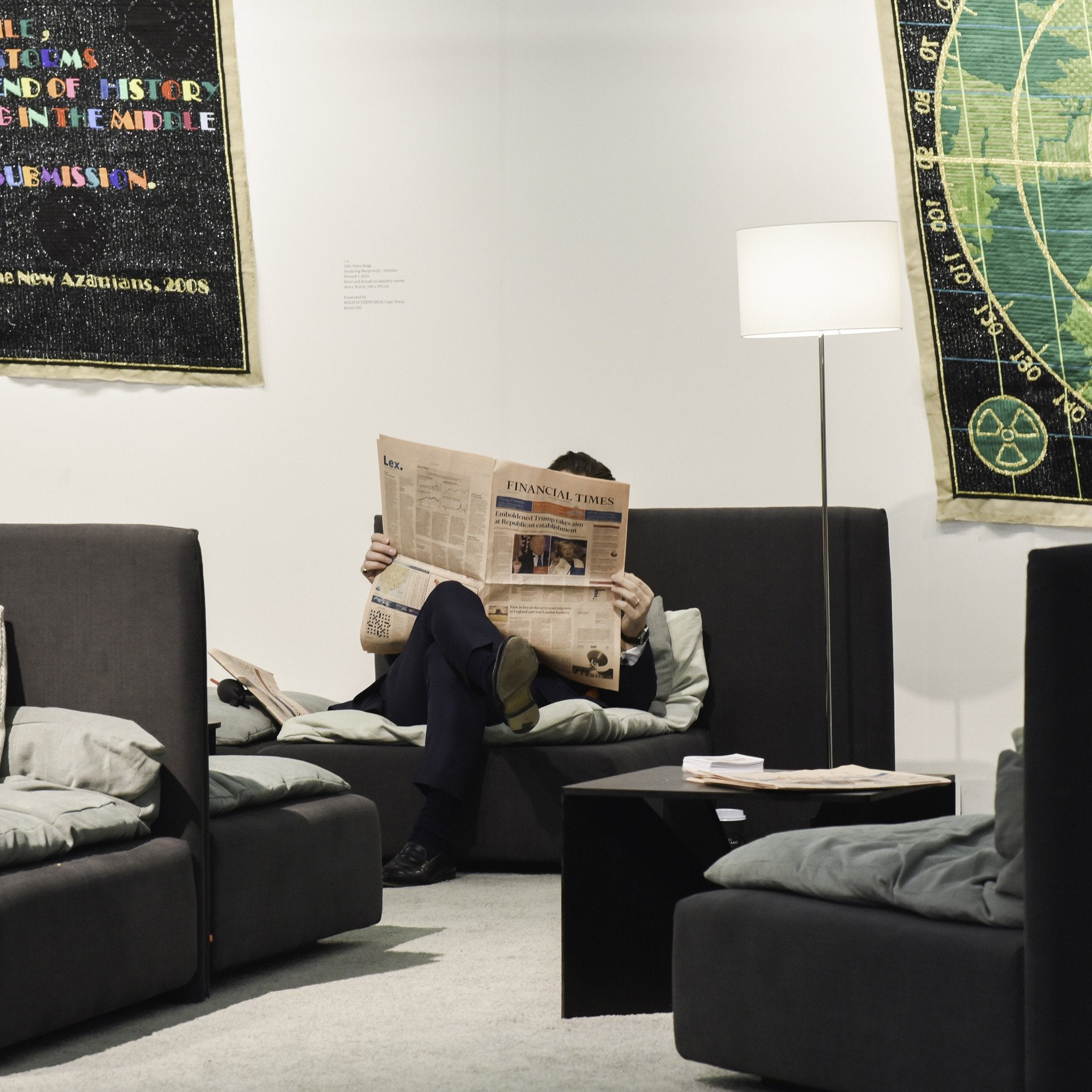Lounging on an FK04 CALVERT sofa by E15, reading the Financial Times beside a chic coffee table. The dark cushions and floor lamp complement the colorful tapestry on white walls, echoing Ferdinand Kramers design style.