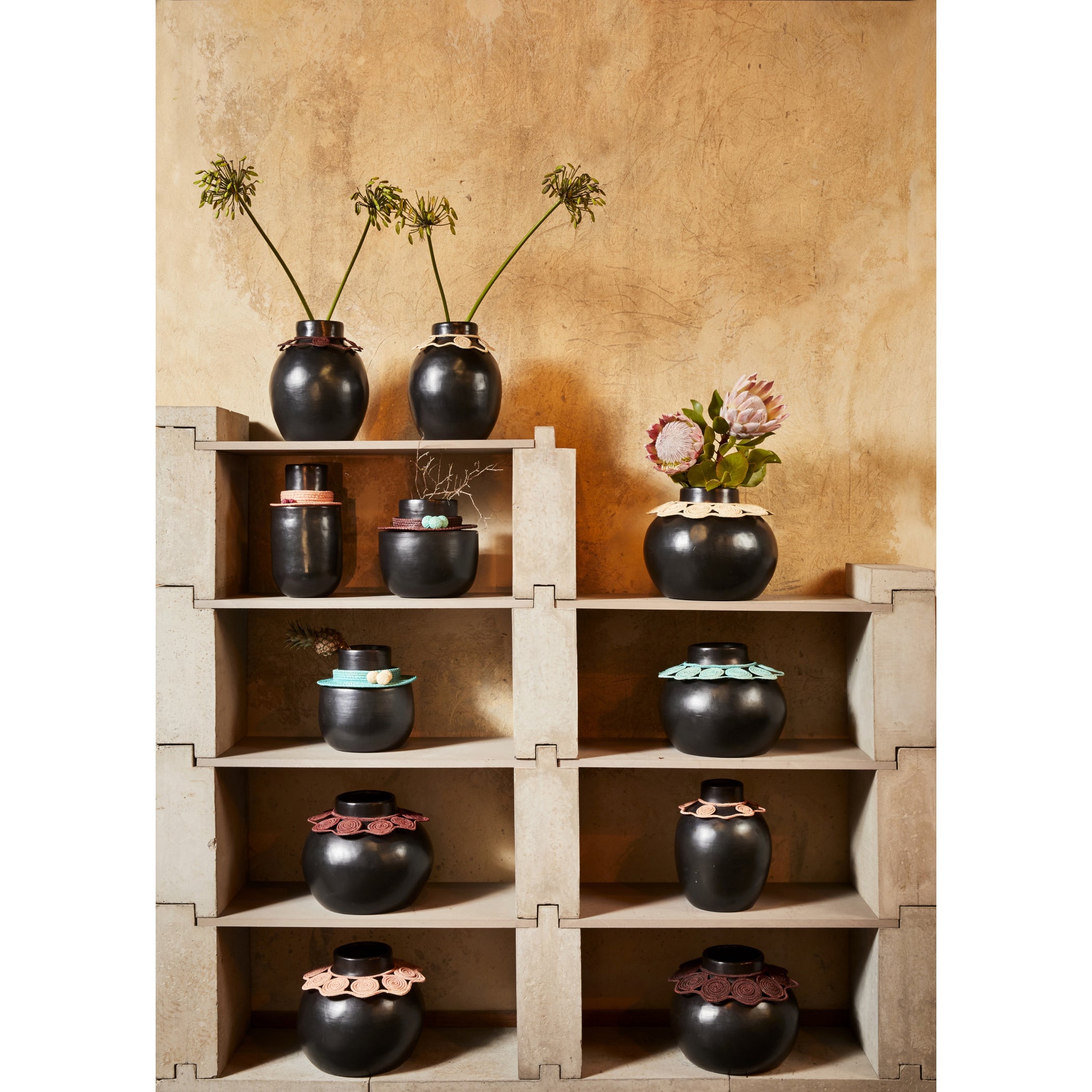 Ames Coyar Vase 1 by Ames, featuring black ceramic with colorful wickerwork and assorted flowers, is displayed on beige stacked shelves against a textured tan wall.
