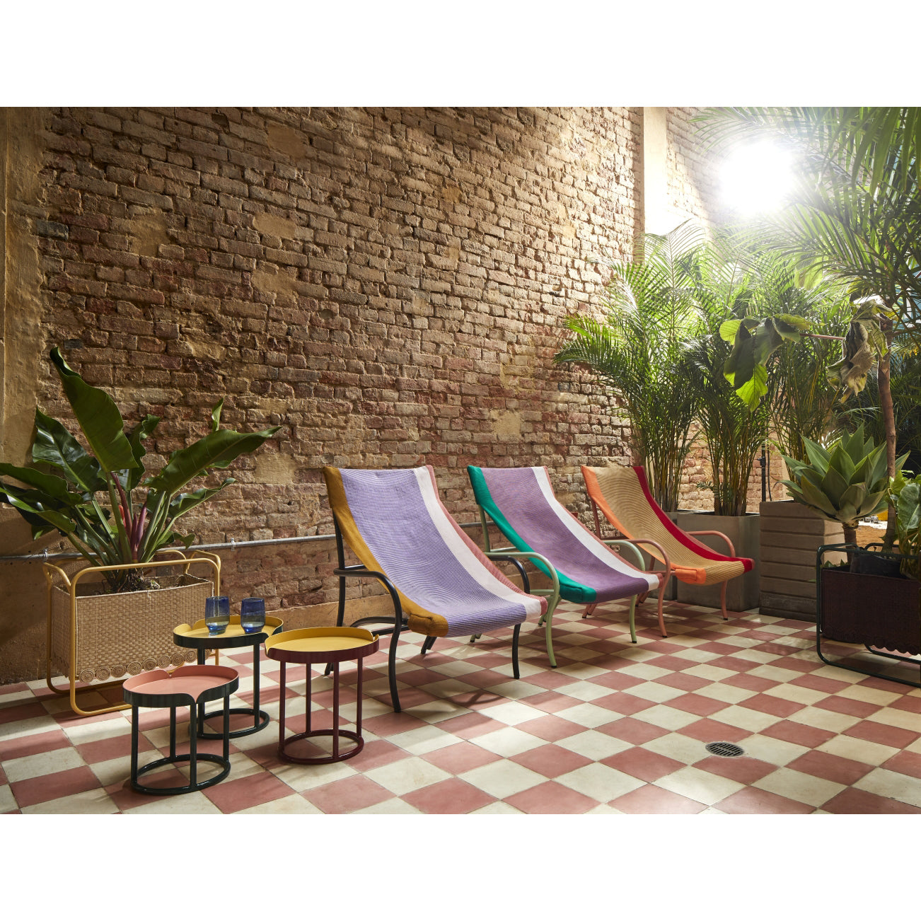 An outdoor patio showcases four vibrant Ames Maraca Lounge Chairs with ethnic designs set against a brick wall, surrounded by potted plants. The floor features a red and white checkered pattern, two small tables hold drinks, and warm lighting enhances the scene.