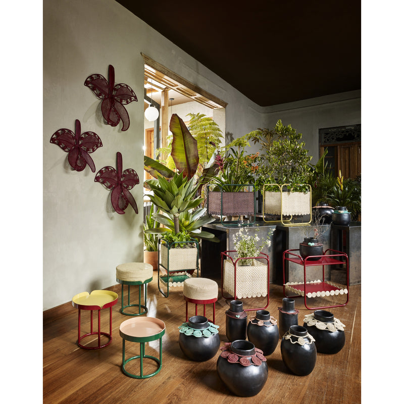 A room decorated with potted plants, colorful stools, black clay vases with decorative rims, and three Ames Cattleia Wall Decor pieces by Ames. Natural light streams in, highlighting the lush greenery and eclectic accents.