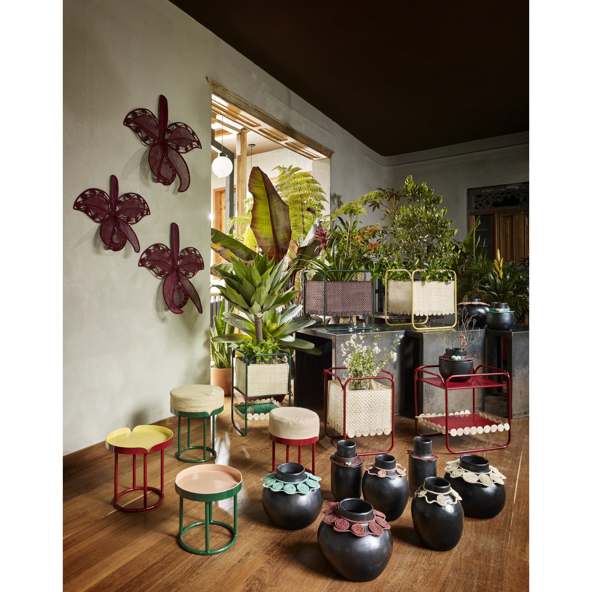 An indoor garden is adorned with diverse plants, including a lush Jipi palm in decorative pots. Red butterfly wall art and colorful Coyar Vase 4 by Ames add artistry, while wickerwork decorations enhance the scene. Sunlight through a window amplifies the natural ambiance.