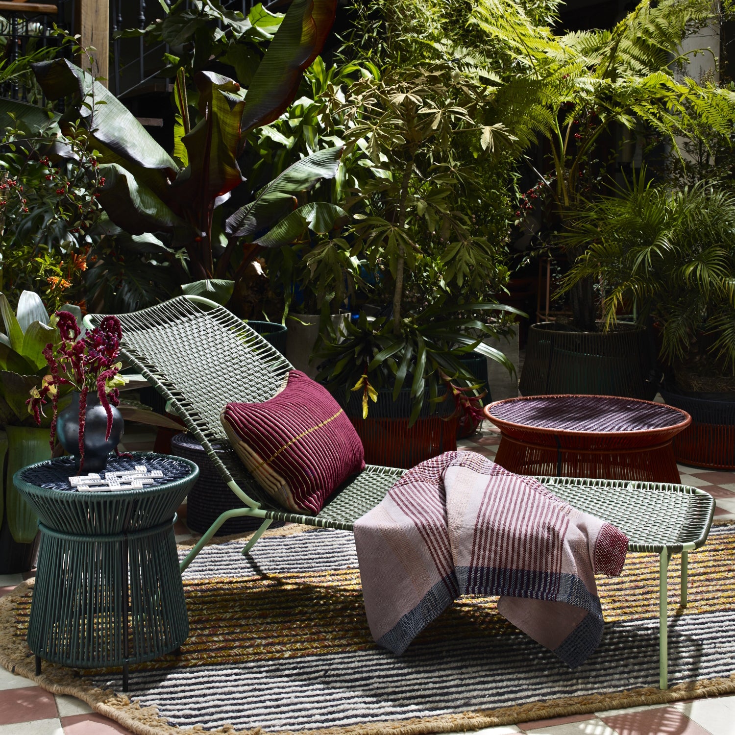 The Ames Cielo Daybed by Ames, topped with a red pillow and striped blanket, rests on a rug amid tropical plants. Two round side tables, one holding a vase, complete this sunlit indoor garden oasis.