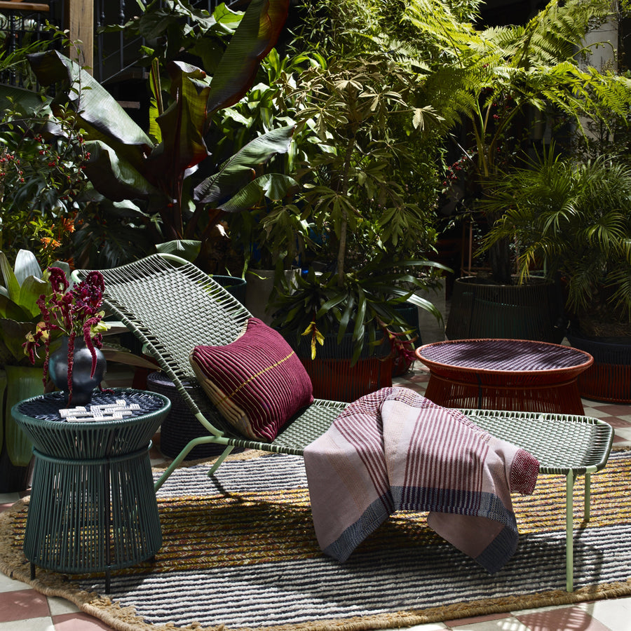 The Ames Cielo Daybed by Ames, topped with a red pillow and striped blanket, rests on a rug amid tropical plants. Two round side tables, one holding a vase, complete this sunlit indoor garden oasis.