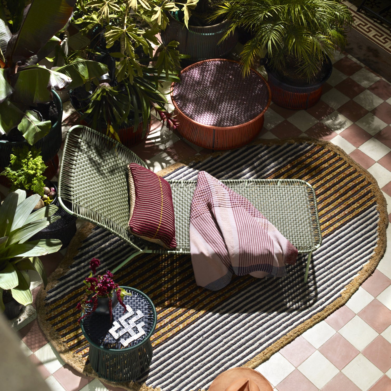A cozy outdoor lounge features a woven chair with striped pillows and a blanket, next to the elegant Ames Cielo Daybed by Ames, surrounded by lush plants, round ottomans, and a patterned rug atop a tiled floor.
