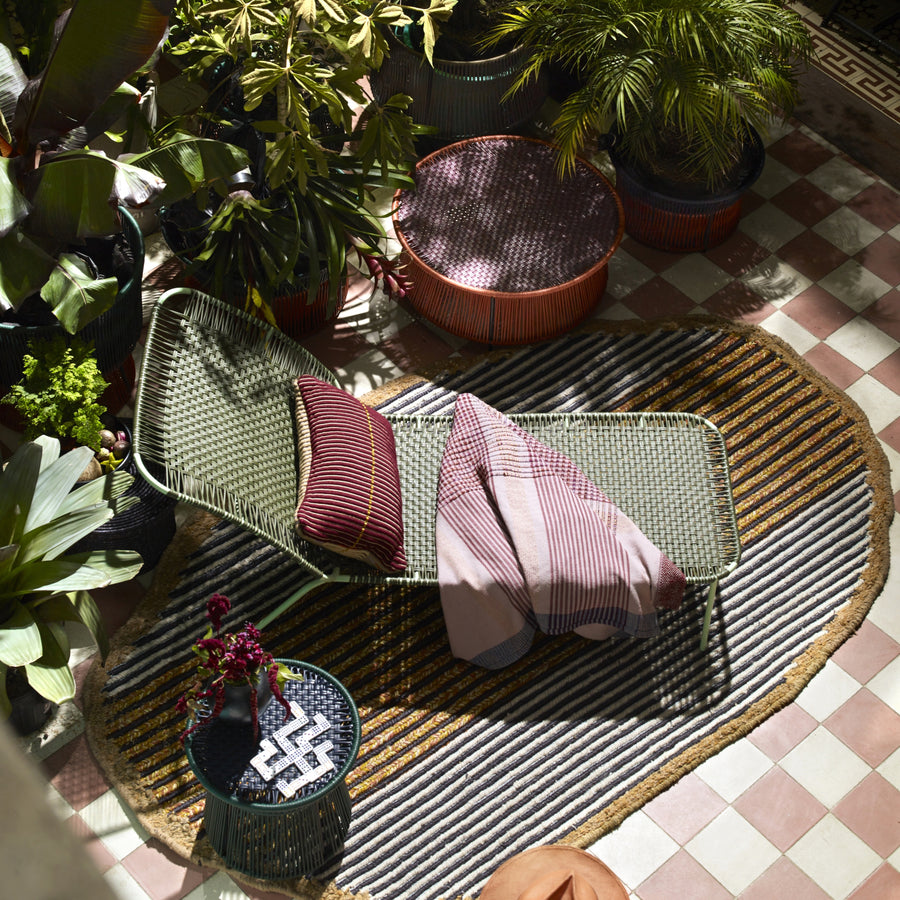 A cozy outdoor lounge features a woven chair with striped pillows and a blanket, next to the elegant Ames Cielo Daybed by Ames, surrounded by lush plants, round ottomans, and a patterned rug atop a tiled floor.
