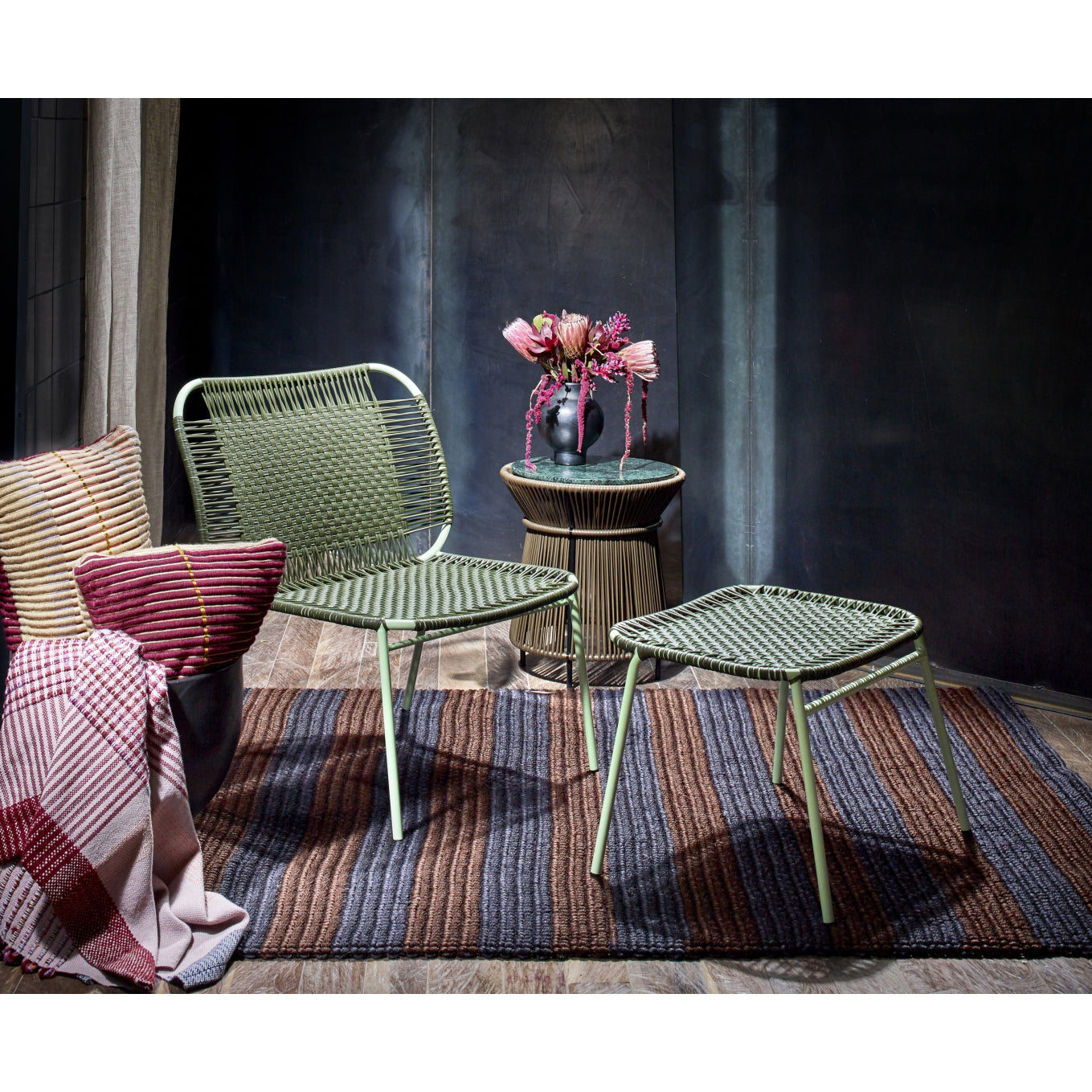 A modern living space featuring the versatile Ames Cielo Lounge Chair Low by Ames for indoor and outdoor use, matched with a footrest, striped rug, side table with pink florals, and striped cushions with a plaid blanket against a dark backdrop.