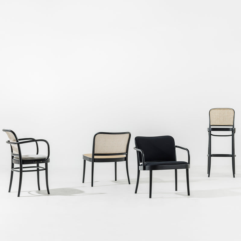 Four TON Lounge Armchairs 811 are set on a plain white background. From left to right: two chairs with wicker backs, a black cushioned chair with elegant upholstery, and a high barstool with a wicker back. All feature dark frames in a minimalist style.