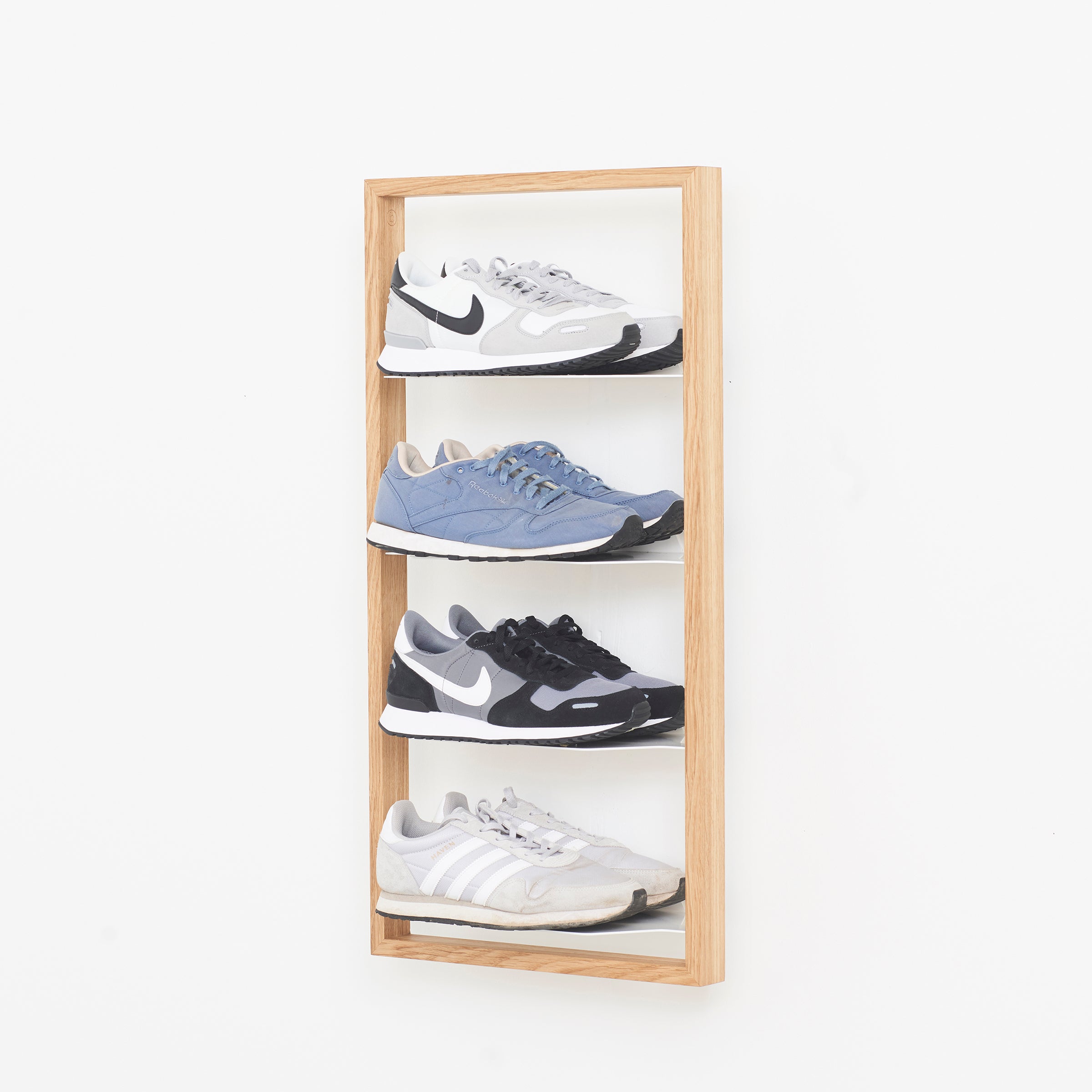 The Basti - Presentation by Das kleine b showcases four sneakers in light gray, blue, black, and white on a wooden wall-mounted rack against a plain white background.