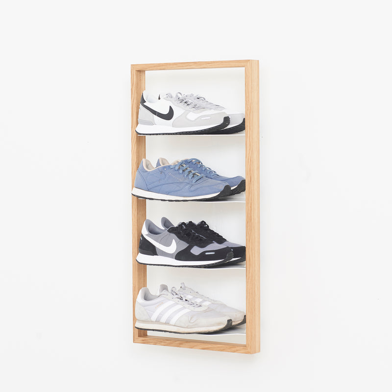 The Basti - Presentation by Das kleine b showcases four sneakers in light gray, blue, black, and white on a wooden wall-mounted rack against a plain white background.