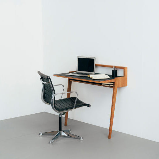 A minimalist office setup features a sleek black chair and solid oak desk against a white wall. The elegantly designed Bachmann Desk by Das kleine b hosts a closed laptop, white notebook, and pen, reflecting a modern workspace thats clean and uncluttered.