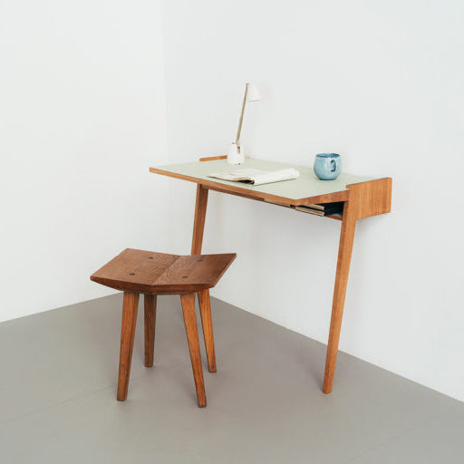 The Bachmann Desk by Das kleine b is a minimalist piece featuring a slanted leg design in solid oak. It includes a matching square stool, topped with an open book, white anglepoise lamp, and blue mug, set against a plain white wall and gray floor.